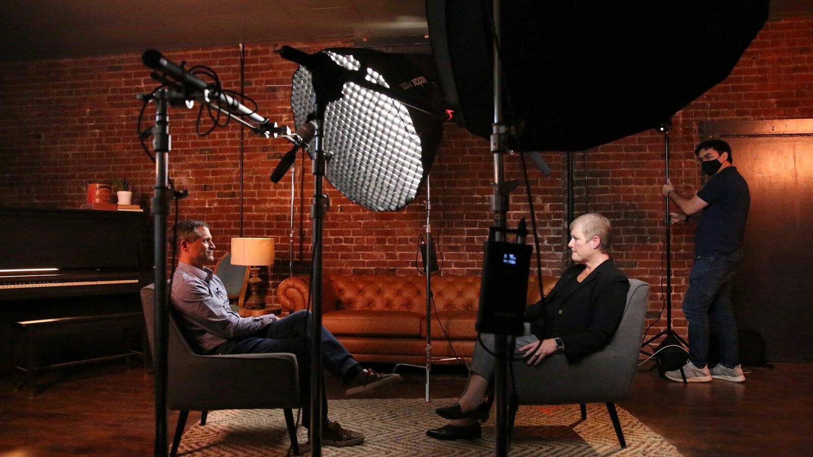 An interview set with a guest and host facing each other under studio lights, microphones and a crew member in the background